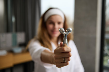Focus on a facial massager with two faceted rollers for rejuvenating facial massage in the hands of a blurred red-haired woman in a white bathrobe during her morning routine in the bathroom at homeの写真素材