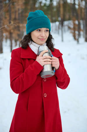 Young beautiful Middle Eastern woman in warm red coat holding a vacuum flask, standing in the snow covered forest, relaxing outdoor on a beautiful cold winter day.の写真素材