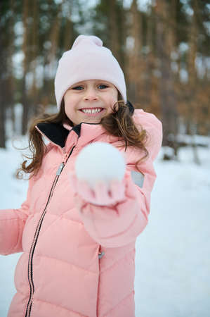Adorable little Caucasian girl in warm pink pastel down jacket showing a snowball, smiling looking...