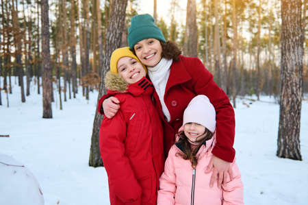 Delighted loving mother- beautiful woman gently hugging her adorable children, boy and girl, smiling with cheerful toothy smile while walking on a snow covered forest path on a winter sunny dayの写真素材