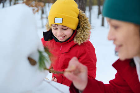 Handsome European boy in bright yellow knit hat making a snowman with his mother and sister in a...
