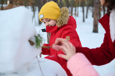Handsome school age boy in bright yellow knit hat making a snowman with his mother and sister in a winter snow covered park. Enjoy wonderful winter outdoor. Leisure activity and happy family pastimeの写真素材
