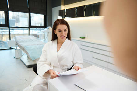 Beautiful European woman holding a checklist while having appointment with beautician doctor, consulting about anti-aging procedures, in beauty clinic with modern equipment for professional body careの写真素材