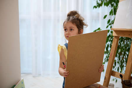 Adorable preschool European girl stands at a wooden easel and looks away while holding a pencil and...