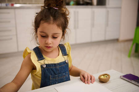 Adorable little European girl in yellow t-shirt and blue denim sitting at table at home and drawing picturesの写真素材