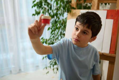 Handsome Caucasian boy in blue t-shirt holding a jar with red gouache paint
