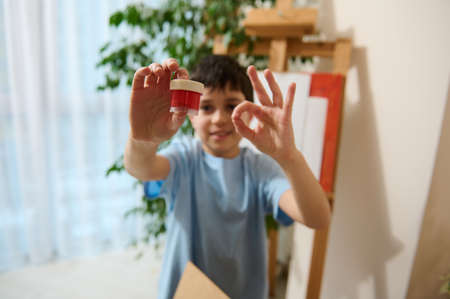 Out of focus dark-haired school boy holding a bottle with red gouache paints and showing OK sign...