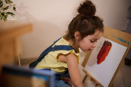 Adorable concentrated little Caucasian girl in yellow t-shirt and blue denim drawing picture at home, using watercolor paints, sitting on the floor in front of wooden easel, enjoying a creative dayの写真素材