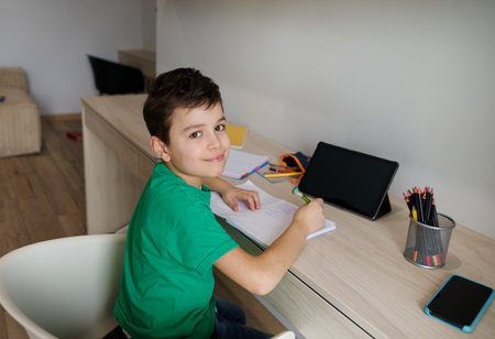Handsome Caucasian 9 years old smart school boy sits at wooden desk in front of a digital gadget and doing his homework, cutely smiles looking at camera. Education and school concept. Online learningの写真素材