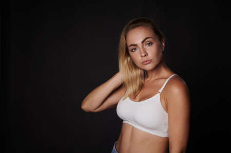 Beautiful young blonde woman with perfect aesthetic body in white sport top confidently looking at camera, isolated over back background with copy ad spaceの写真素材