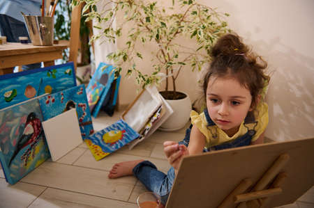 Beautiful European little girl enjoying learning painting, sitting on the floor at a wooden easel against the background of colorful joyful children painted images. Art class, creativity and educationの写真素材