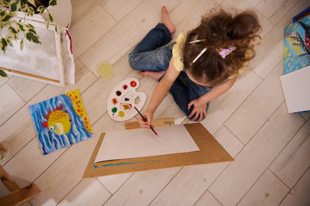Overhead view cute European little girl enjoying painting, sitting on the floor at a wooden easel against the background of colorful joyful children painted images. Art class, creativity and educationの写真素材
