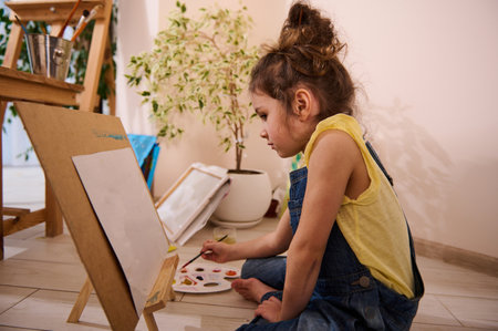 Adorable European child, cute little girl sitting on the floor at a wooden easel and drawing picture painting on canvas. Art class, creativity, children entertainment concept.の写真素材