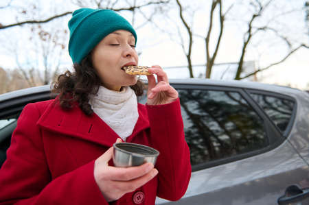 Beautiful Caucasian woman, female driver traveler eating take out food, delicious baked cookies, and drinking a hot tea from a thermo mug, standing near her car in the snowy forest . Wonderful winterの写真素材