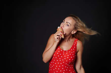 Attractive stunning blonde Caucasian woman with flying hair from the wind, wearing a red swimsuit with white polka dots eats ice cream cone, looks at camera posing against black background, copy spaceの写真素材