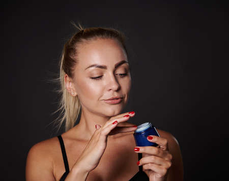Headshot stunning beautiful middle aged European blonde woman applying face moisturizer, smoothing...