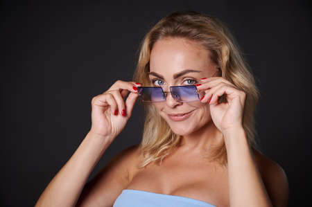 Studio portrait beautiful blonde Caucasian woman confidently looking at camera through blue sunglasses, isolated over black background with copy space for adsの写真素材