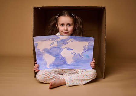 Adorable Caucasian child 4 years old girl sitting inside a cardboard box with a poster- painted image with World map, isolated over beige background.Environment conservation and save planet conceptの写真素材