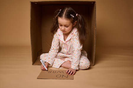 Cute child, adorable Caucasian kid girl in home clothes hiding from war inside cardboard box, writing social message- Stop War. Concepts of refugees migrants losing their home during military conflictの写真素材