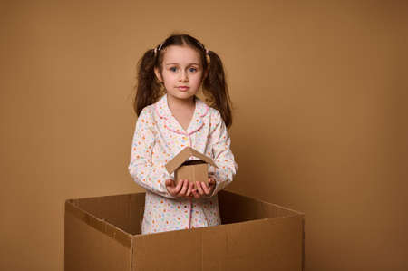Little Caucasian girl, adorable child in pajamas holds a craft cardboard house model being inside a box, against beige background with copy space for ads. The concept of investment, housingの写真素材