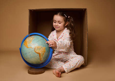 Adorable child baby girl in pajamas sitting ahead a cardboard box and examining the Earth globe, isolated over beige background with copy space. Love and save planet, environment conservation conceptの写真素材