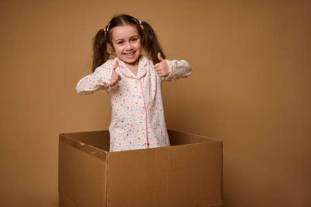 Adorable Caucasian little girl, cheerful kid with two ponytails showing thumbs up, smiles looking at camera being inside a cardboard box against a beige background with copy advertisement spaceの写真素材