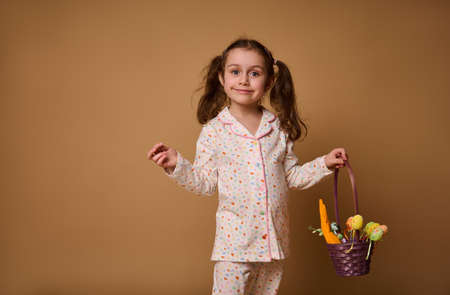 Portrait of a beautiful Caucasian child girl in pajamas with bright dots stands with a purple basket and colored Easter eggs, isolated on a beige background with space for advertising textの写真素材