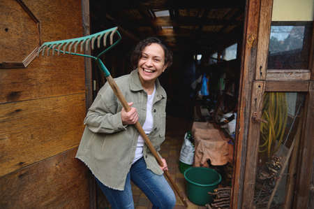 Happy pleasant mature female gardener holding a rake and smiling with a cheerful toothy smile while...