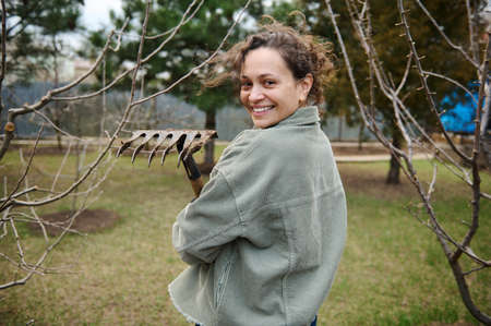 Portrait of a female gardener, beautiful woman holding a soiled rake in her hands and smiles looking...