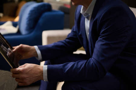 Cropped image. Hands of a man in business suit holding a catalog and sitting on a sofa in the showroom of an upholstered furniture store. Small, business, interior design, home improvement conceptの写真素材