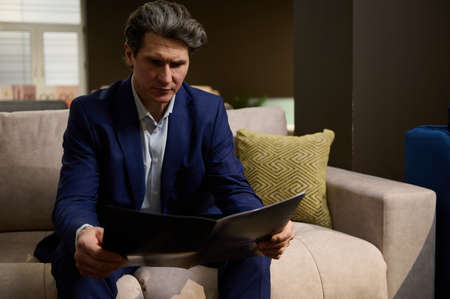 Handsome imposing mature Caucasian man in business suit looking at catalog with upholstered sofas and beds while visiting a furniture store showroomの写真素材