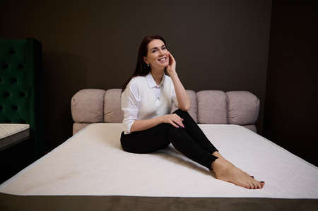 Attractive Caucasian pretty woman in casual attire sitting on an orthopedic mattress in the furniture store. The choice of mattresses in the exhibition center of a furniture storeの写真素材