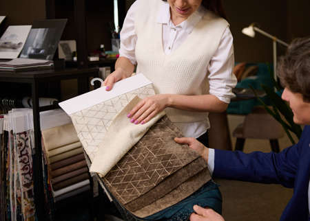 Close-up of a sales assistant showing fabric samples for sofa upholstery to customer in furniture store. Home decoration repair upholstery planning.の写真素材