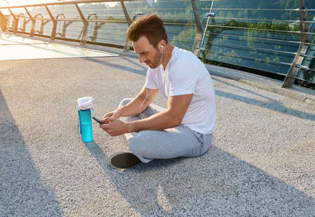 Middle aged handsome Caucasian sportsman wearing earphones sitting on the city bridge and checking...