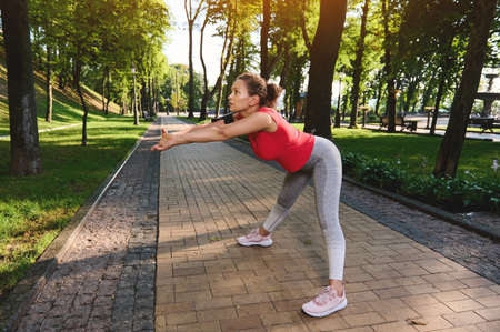 Charming middle aged sportswoman in tight sportswear warming up and stretching her body, doing...