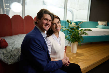 Charming middle-aged European couple in furniture store, shopping for soft bed and sofa, holding hands and smiling looking at camera. Home improvement and interior design conceptの写真素材