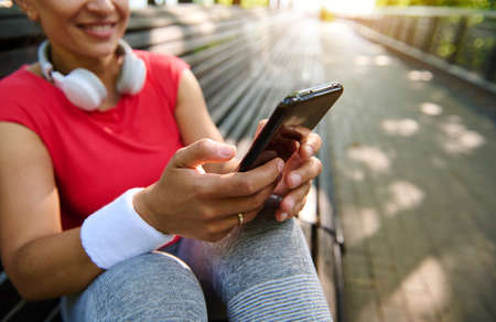 Focus on the hands with smartphone of athletic woman with headphones tapping and texting message on sunny warm summer day. Beautiful cheerful young sportswoman using phone for chatting after workoutの写真素材
