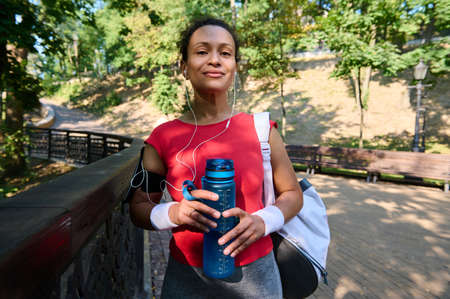Confident African American athletic woman in red tight t-shirt and terry wristbands holds bottle...