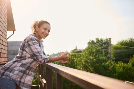 Beautiful delightful young Hispanic curly haired woman in casual clothes drinks coffee and enjoys the view of the country houses from the balcony on a beautiful warm sunny dayの写真素材