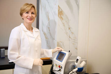 Experienced charming Caucasian mature female doctor in white medical gown and sterile surgical gloves , smiles at camera standing near ultrasound medical equipment in doctor's cabinetの写真素材