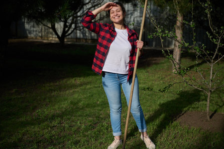 Happy female gardener standing in the backyard with a gardening tool in her hand and smiling toothy...