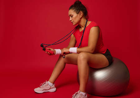 Attractive determined fit woman in sportswear sits on a fit ball with a skipping rope in her hands,...