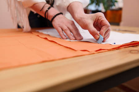 Tailor at work. Close-up of a dressmaker's hands transferring sewing patterns for clothes on orange colored satin fabric in a clothing fashion design studioの写真素材
