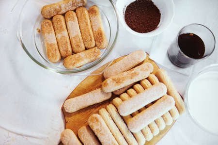 Flat lay. Ingredients for making traditional Italian tiramisu on kitchen table - savoiardi biscuit cookies on wooden board, sweet espresso coffee in glass, mascarpone cheese and bowl with fresh creamの写真素材
