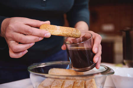 Details: confectioner hands dipping ladyfinger biscuit cookies into a glass of strong sweet espresso coffee. Step-by-step recipe for making traditional Italian dessert Tiramisu at homeの写真素材