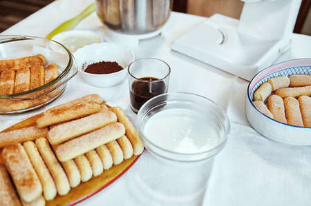 Ingredients for making the traditional Italian sweet tiramisu cake. Savoiardi cookies, strong sweet espresso coffee, cocoa powder, mascarpone cheese and fresh cream on a table with a white tableclothの写真素材