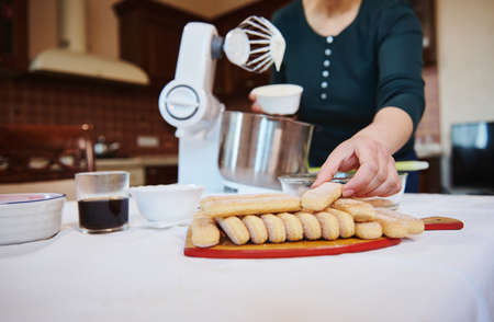 Details of a woman hands holding sponge biscuits ladyfingers while preparing homemade Italian dessert Tiramisu in a cozy home wooden kitchen. Step by step recipe. Homemade food, culinary concept.の写真素材