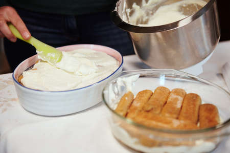 Close-up. Chef's hand putting mixed mascarpone and whipped cream cheese cream on the cake while making tiramisu - traditional Italian dessert at home. Step-by-step recipeの写真素材