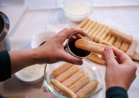 Details of a confectioner hands dipping ladyfinger biscuit cookies into a glass of strong sweet espresso coffee. Step-by-step recipe for making traditional Italian dessert Tiramisu at home. Culinaryの写真素材