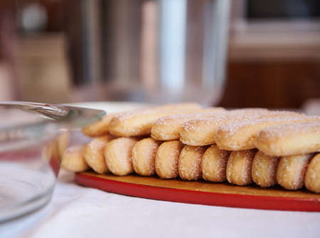 Close-up of ladyfinger savoiardi biscuit cookies on a wooden cutting board on white tablecloth background with blurred steel bowl and glass plate. Close upの写真素材